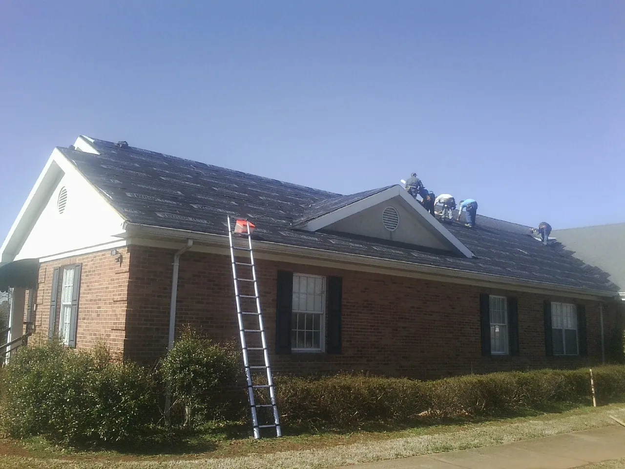 Crew tearing shingles off a brick ranch roof with a ladder set — Emergency Roof Repair work by our crew in Bishop Hill