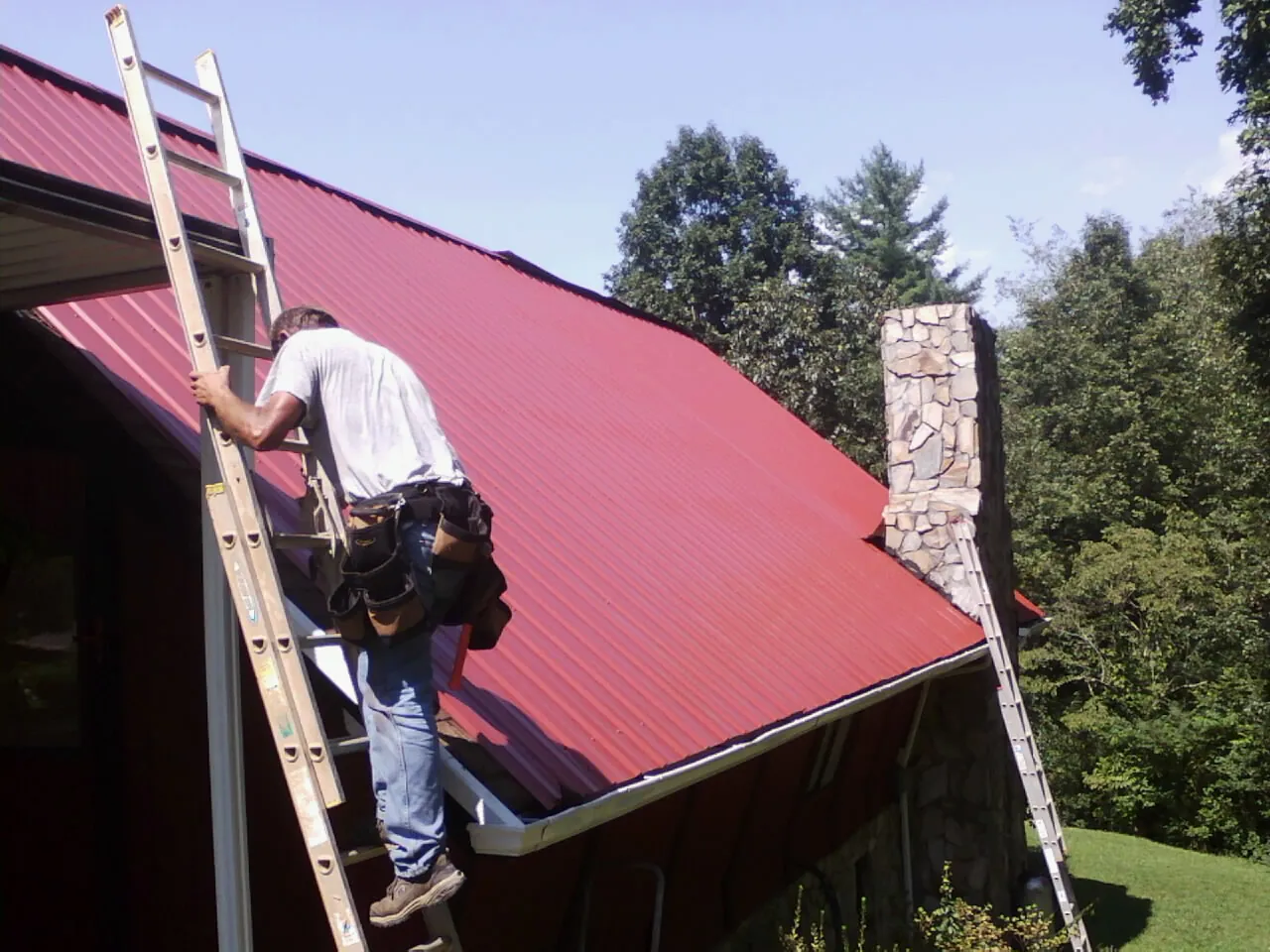 Roofer on a ladder beside a red metal roof and stone chimney — Soffit & Fascia Repair by Roofing Brothers in Bishop Hill